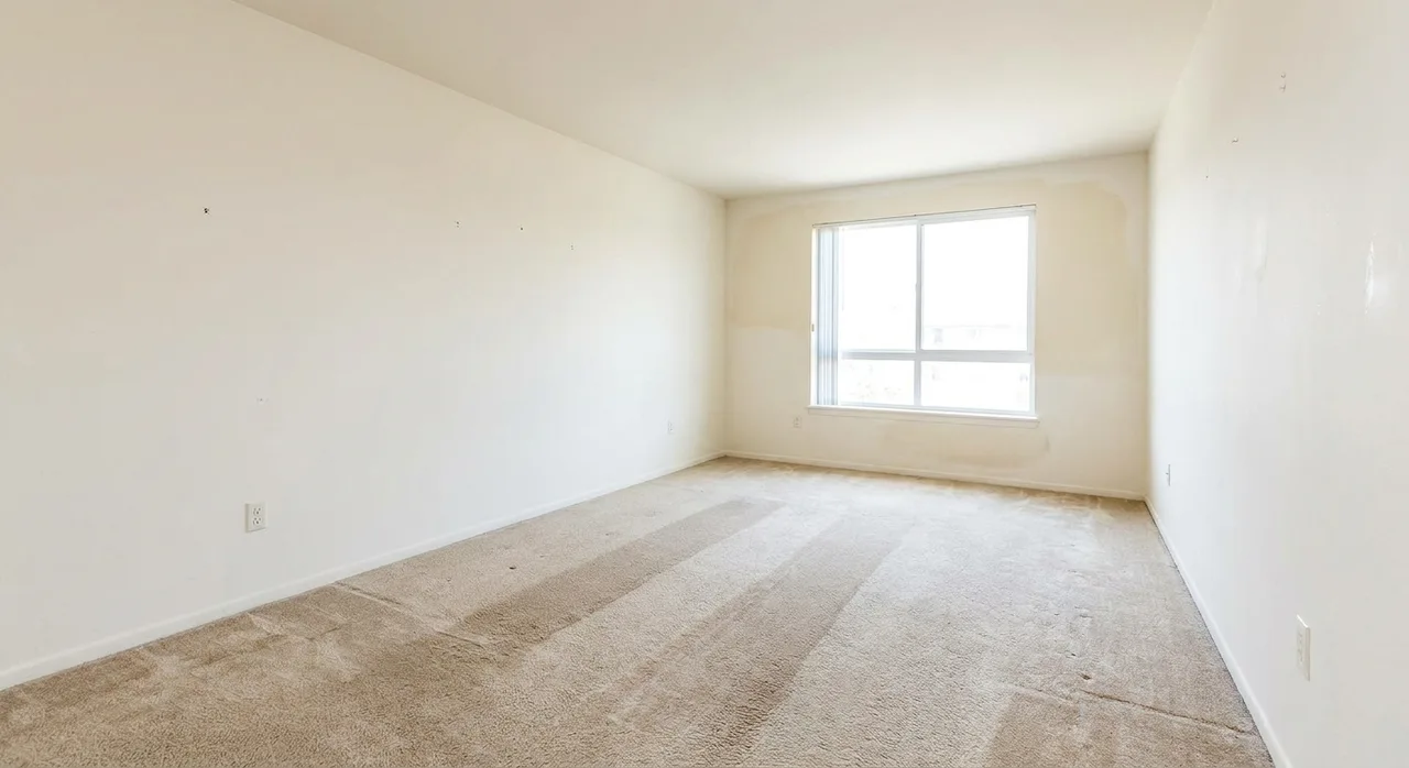 A normally worn unit and a damaged one look different, and the difference decides the deposit An empty rental living room in good condition showing normal wear: worn carpet traffic paths and lightly scuffed walls